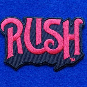 May include: An embroidered patch featuring the word RUSH in bright pink thread with a black border. The letters are stylised with a bold, curved font. The patch is shaped to follow the outline of the text, creating a distinct graphic emblem.