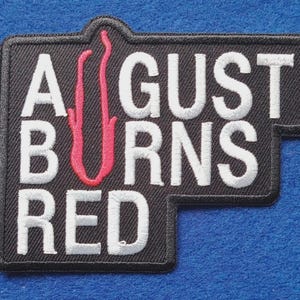 May include: A black embroidered patch with white text that reads AUGUST BURNS RED. The letter U in AUGUST is replaced by a stylised red flame graphic. The patch has a rectangular shape with a stepped edge on the right side and black stitching.
