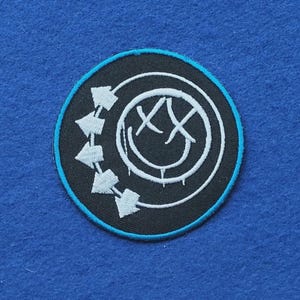 May include: A circular embroidered patch featuring a black background with a bright blue border. The design includes a white smiley face with X-shaped eyes and a dripping mouth, surrounded by a partial ring of five white arrows pointing clockwise.