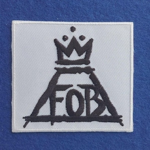 Puede incluir: Un parche cuadrado bordado en blanco que presenta un diseño cosido en negro de un triángulo que contiene las letras FOB, rematado con un gráfico de corona que consta de tres puntas y tres puntos pequeños sobre el pico central.