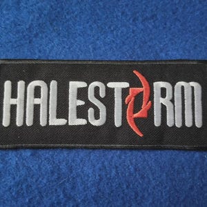 May include: A rectangular black embroidered patch featuring the word HALESTORM in white capital letters. The letter O is replaced by a stylised red symbol consisting of two curved, pointed shapes facing each other with a black centre.