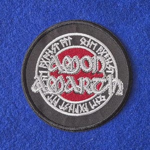 May include: A circular embroidered patch featuring a black border and a red centre circle. The text Amon Amarth is stitched in white in the middle, surrounded by a ring of white runic-style lettering on a black background.