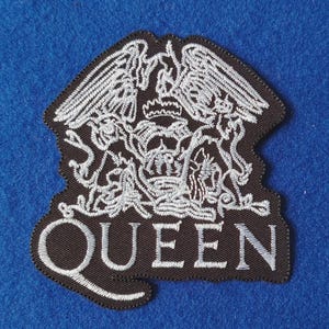 Op de afbeelding: Een zwarte geborduurde patch met het Queen-bandlogo in wit garen. Het ontwerp bevat een centraal wapenschild met mythische wezens, een kroon en het woord QUEEN in hoofdletters onderaan, alles omlijnd door een donkere rand.