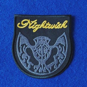 May include: A shield-shaped embroidered patch with a black background. The word Nightwish is stitched in yellow cursive script at the top. Below the text, a grey, intricate, symmetrical emblem resembling a stylised thistle or crest is featured.