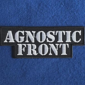 Può includere: Una toppa ricamata rettangolare con sfondo nero e bordo bianco. Il testo AGNOSTIC FRONT è ricamato in lettere maiuscole bianche al centro, con la parola AGNOSTIC sulla riga superiore e FRONT sulla riga inferiore.