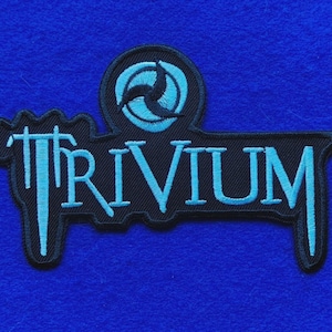 May include: A black embroidered patch featuring the word TRIVIUM in a stylised light blue font. Above the text is a circular emblem containing a light blue three-pointed symbol on a black background. The patch has a black border and edges.