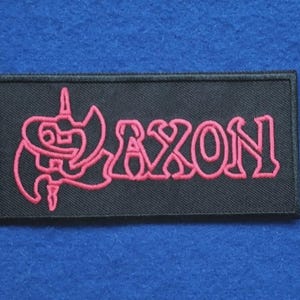 Peut inclure: Un écusson rectangulaire noir brodé avec le mot AXON en fil rose vif. À gauche du texte se trouve un symbole graphique rose stylisé ressemblant à une hache à double tranchant ou à un blason. L'écusson possède une bordure noire cousue.