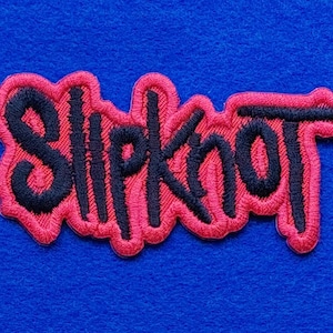 Peut inclure: Un écusson brodé représentant le mot Slipknot en fil noir avec une bordure rouge épaisse et texturée. Le lettrage est stylisé dans une police manuscrite dentelée, et l'ensemble du motif est souligné en rouge, créant un contraste marqué.