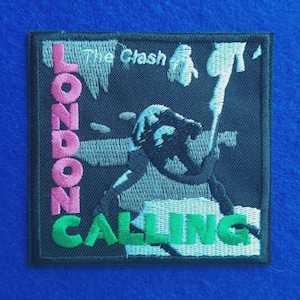 May include: A square embroidered patch featuring a black and white graphic of a person smashing a guitar. The text "The Clash" appears in white, "LONDON" in pink, and "CALLING" in green. The patch has a black border and a dark background.