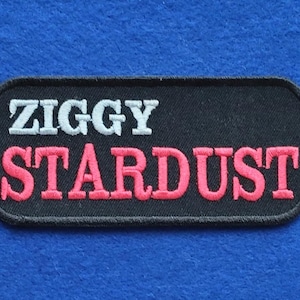 Könnte beinhalten: Ein rechteckiger bestickter Aufnäher mit schwarzem Hintergrund und abgerundeten Ecken. Der Text ZIGGY ist in weißem Garn in der oberen Zeile gestickt, und das Wort STARDUST ist in leuchtend rosa Garn in der unteren Zeile in Serifenschrift.