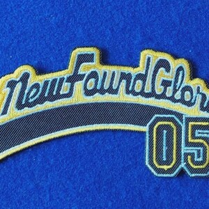 May include: An embroidered patch featuring the text New Found Glory in a stylized black font with a light blue outline and a yellow border. Below the text, the number 05 is displayed in a matching black, light blue, and yellow design.