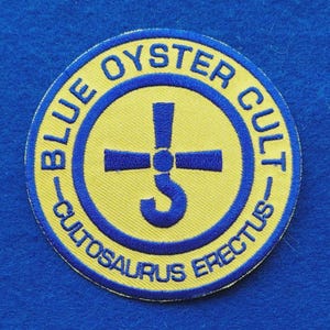 Può includere: Una toppa ricamata circolare con sfondo giallo e bordo blu. Il testo BLUE OYSTER CULT e CULTOSAURUS ERECTUS è ricamato in blu. Un simbolo blu che ricorda una croce con un gancio nella parte inferiore è centrato sulla toppa.