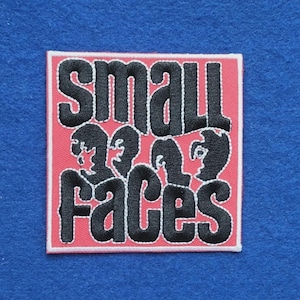 Puede incluir: Un parche bordado cuadrado con fondo rosa y borde blanco. El texto "SMALL FACES" está bordado en letras negras gruesas, con siluetas negras estilizadas de rostros integradas en el diseño de las letras.