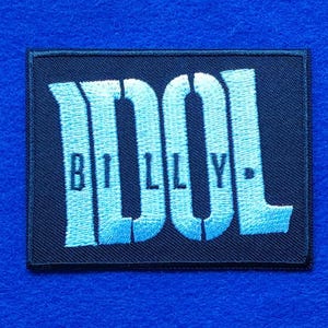 May include: A rectangular black embroidered patch featuring the word IDOL in large, light blue, sans-serif capital letters. The word BILLY is stitched in smaller, dark blue, serif capital letters across the centre of the larger text.