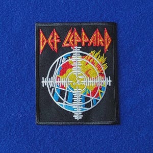 May include: A rectangular embroidered patch with a black background featuring the text DEF LEPPARD in red and yellow letters. A white crosshair graphic overlays a yellow and red flame design with blue accents, all bordered by a black stitched edge.