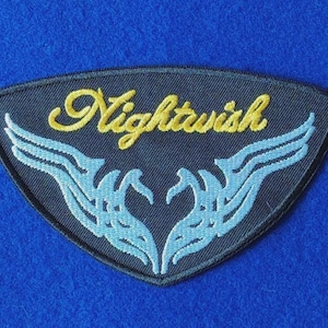 May include: A shield-shaped embroidered patch with a black background. The word Nightwish is stitched in yellow cursive script above a symmetrical light blue graphic design that resembles stylized wings or abstract flame patterns.