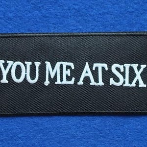 Puede incluir: Un parche rectangular bordado en negro con el texto blanco YOU ME AT SIX en una fuente serif negrita. El parche tiene un borde cosido en negro y una superficie de tela texturizada, diseñado para su aplicación en ropa u otros textiles.