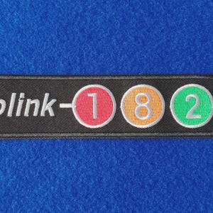 May include: A rectangular black embroidered patch featuring the white text blink- followed by three circular icons. The first circle is red with a white number 1, the second is orange with a white number 8, and the third is green with a white number 2.