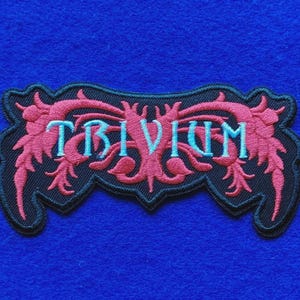 Op de afbeelding: Een zwarte geborduurde patch met het woord TRIVIUM in lichtblauwe hoofdletters. De tekst is omringd door complexe, symmetrische roze bloem- of rankpatronen, allemaal omlijnd met een zwarte rand op een donkere stoffen ondergrond.