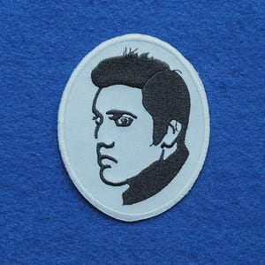 May include: An oval-shaped embroidered patch featuring a black thread portrait of a person with a stylised hairstyle on a light blue background. The design shows a side profile of a face with dark hair, defined eyebrows, and a serious expression.