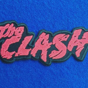 Op de afbeelding: Een geborduurde stoffen patch met de tekst The Clash in een gestileerd, grillig rood lettertype. De letters zijn omlijnd met een dikke zwarte rand, wat een duidelijk contrast vormt met het rode borduurwerk. De patch heeft een textuurrijk oppervlak.