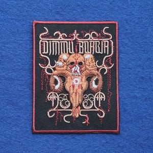Op de afbeelding: Een rechthoekige geborduurde patch met een zwarte achtergrond en een rode rand. Het ontwerp bevat de tekst DIMMU BORGIR in een gestileerd lettertype boven een beige afbeelding van een gehoornde schedel met rode en witte draaddetails.