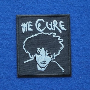 Könnte beinhalten: Ein rechteckiger, schwarz bestickter Aufnäher mit dem weißen Schriftzug The Cure im oberen Bereich. Darunter befindet sich eine weiße Linienzeichnung einer Person mit voluminösem Haar und markanten Gesichtszügen, eingefasst von einem Rand.