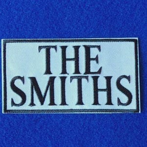 Peut inclure: Un écusson rectangulaire brodé présentant un fond blanc avec une bordure noire et le texte THE SMITHS en lettres capitales noires en gras. Le motif est centré sur le tissu, mettant en valeur des coutures nettes et un lettrage contrasté.