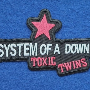 Könnte beinhalten: Ein schwarzer gestickter Aufnäher mit einem rosa Stern an der Oberseite. Der Text SYSTEM OF A DOWN ist in Weiß gestickt, während die Wörter TOXIC TWINS darunter in Rosa gestickt sind. Das gesamte Design ist mit einem schwarzen Rand versehen.