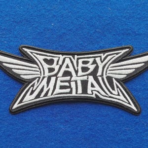 Può includere: Una toppa ricamata in bianco e nero con il testo BABY METAL in un carattere stilizzato. Il design include due ali simmetriche che si estendono dai lati della scritta centrale, il tutto delineato da un bordo nero spesso con bordi netti.