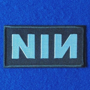 May include: A rectangular embroidered patch featuring a black background with the letters NIN stitched in light blue thread. The letter N is mirrored on the right side, creating a distinct logo design with a clean, stitched border edge.