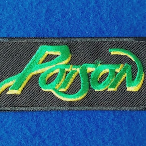May include: A rectangular embroidered patch featuring a black background with the word Poison stitched in a stylised green font with a yellow outline. The embroidery has a textured appearance with a thick black border surrounding the entire design.
