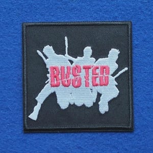 Op de afbeelding: Een vierkante zwarte geborduurde patch met een witte silhouet van drie figuren die muziekinstrumenten bespelen. Het woord BUSTED is in felroze hoofdletters over het midden van het witte silhouetontwerp geborduurd.