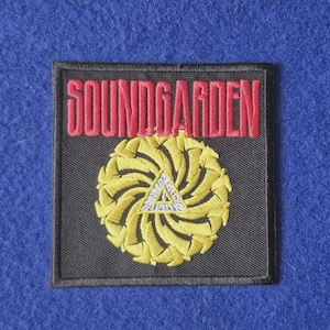 May include: A square black embroidered patch featuring the red text SOUNDGARDEN at the top. Below the text is a circular yellow design composed of swirling shapes surrounding a small white triangle with a central eye symbol.