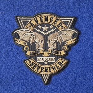 May include: A black and tan embroidered patch featuring the text Avenged Sevenfold, Orange County, and California. The design includes two winged skulls, lightning bolts, and stars arranged within a triangular border with a black background.
