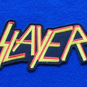 Peut inclure: Un écusson brodé rectangulaire présentant le mot SLAYER dans une police stylisée. Les lettres sont cousues avec un fil jaune vif avec un contour rouge audacieux, le tout sur un fond noir uni avec une bordure noire épaisse.