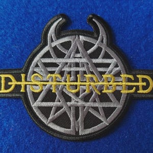 May include: An embroidered patch featuring the word DISTURBED in yellow thread across a circular black and grey geometric design. The emblem includes two curved horn-like shapes at the top and a horizontal line running through the centre text.