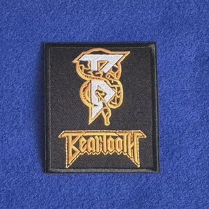 Puede incluir: Un parche rectangular negro bordado que presenta un logotipo estilizado en blanco y dorado de las letras BT entrelazadas con un diseño de serpiente. Debajo del logotipo, la palabra Beartooth está bordada en letras doradas.