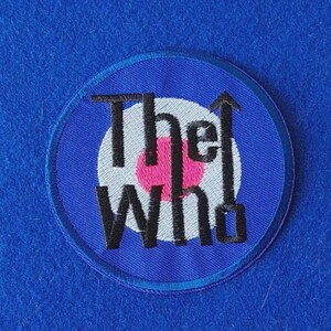 Può includere: Una toppa ricamata circolare che presenta il testo The Who in lettere nere. Lo sfondo della toppa mostra un design a bersaglio con un cerchio centrale rosa, un anello bianco e un bordo esterno blu, sormontato da un simbolo di freccia nera.