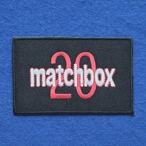 May include: A rectangular black embroidered patch featuring the word matchbox in white lowercase letters overlaid with the large red number 20. The patch has a black stitched border and a textured fabric surface throughout the design.