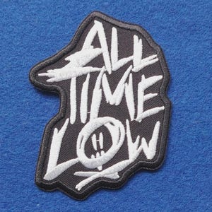 May include: A black embroidered patch with white stitching featuring the text ALL TIME LOW in a stylised font, with the letter O replaced by a circular smiley face icon with a small cross underneath the design.
