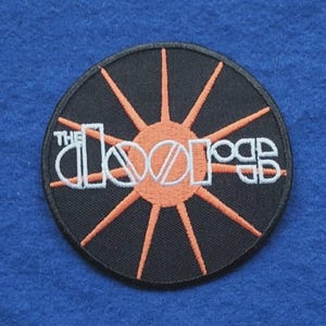 May include: A circular embroidered patch featuring a black background with an orange sunburst design. The white text reads THE DOORS in a stylised font across the centre. The patch has a black stitched border and a flat, circular shape.