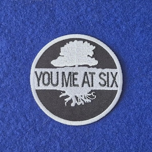 May include: A circular embroidered patch featuring a black background with a white border. The design displays a white tree silhouette with visible roots and the text YOU ME AT SIX printed in white across the centre of the tree graphic.