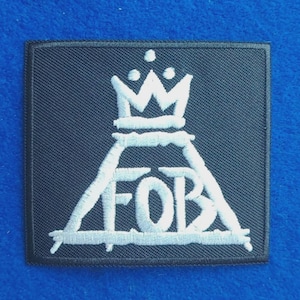 Puede incluir: Un parche cuadrado de tela negra con bordado blanco. El diseño consiste en una forma triangular que contiene las letras FOB, rematada con un gráfico de corona con tres pequeños puntos encima. Los bordes del parche están rematados.