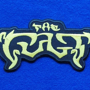 May include: An embroidered patch featuring a black background with the text THE CULT stitched in a stylised, pale yellow font. The lettering has a jagged, decorative appearance with elongated, curved edges that extend across the patch surface.