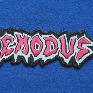 May include: An embroidered patch featuring the word EXODUS in a stylised, jagged font. The lettering is pink with a thick white outline, set against a black background with a rough, irregular border that follows the shape of the text.