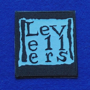 May include: A square embroidered patch featuring a light blue centre with a jagged black border. The text "Levellers" is stitched in black, with "Lev" on the top line, "e11" on the middle line, and "ers" on the bottom line of the patch.