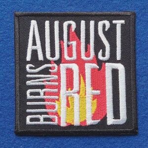 May include: A square embroidered patch with a black background featuring the white text AUGUST BURNS RED arranged in a stacked layout. Behind the text is a stylised flame graphic in shades of red and yellow, all enclosed by a black border.