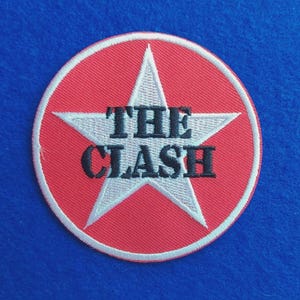 Op de afbeelding: Een ronde geborduurde patch met een rode achtergrond en een witte ster in het midden. De woorden THE CLASH zijn in zwarte hoofdletters over het midden van de ster geborduurd. De patch heeft een witte rand langs de buitenkant.