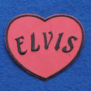 May include: A heart-shaped embroidered patch featuring a vibrant pink background with a black stitched border. The name ELVIS is embroidered in the centre in bold, black, stylised capital letters, creating a clear and distinct visual contrast.